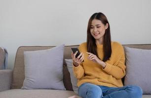 Happy asian woman using mobile phone. Smiling young female using app shopping online, ordering delivery relax on sofa at home. photo