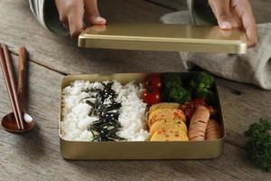 Female Hand Preparing Dosirak or Korean Bento Box, Served on Gold  Metal Lunchbox to Keep Food Warm. photo