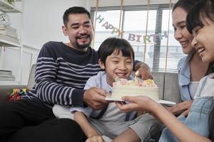 Happy Asian Thai family, young son is surprised with birthday cake, blows out candle, and celebrates joy party with parents and sister in living room together, wellbeing domestic home special event. photo