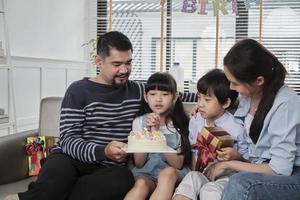 Happy Asian Thai family, young daughter is surprised with birthday cake and gift, blows candle, pray and cheerful celebrates party with parents together in living room, domestic home event lifestyle. photo