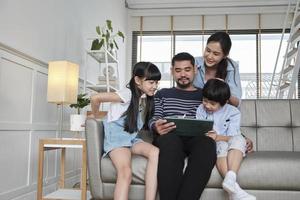 Happy Asian Thai family, parents, and children have fun using digital tablet together on sofa in home living room, a lovely leisure weekend, and domestic wellbeing lifestyle with internet technology. photo