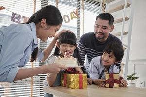 Happy Asian Thai family, young daughter is surprised, birthday cake and lovely gift, girl blows candle, pray and cheerful celebrates party with parents together, special domestic home event lifestyle. photo