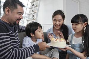 feliz familia asiática tailandesa, el hijo pequeño se sorprende con un pastel de cumpleaños, sopla una vela y celebra la fiesta de alegría con los padres y la hermana en la sala de estar juntos, evento especial de bienestar doméstico en el hogar. foto