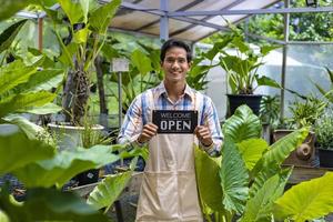 jardinero asiático sosteniendo un cartel de apertura para dar la bienvenida al cliente a su centro de plantas de vivero tropical lleno de especies exóticas de helechos foto