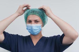 Close up of female doctor or scientist with a medical mask and surgical cap over grey background. She is adjusting mask with photo