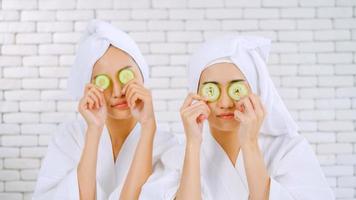 Two happy Asian girls in white bathrobes with towels on heads holding cucumber slices in living room. photo