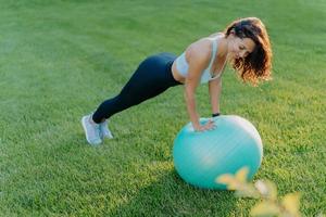 Sporty girl stands in plank pose on gymnastic ball, dressed in sportswear, does physical exercises on green lawn outdoor, keeps fit and healthy. People, yoga, flexibility, lifestyle concept. photo