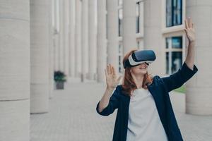 Young ginger woman dressed formally stands outdoors and using VR headset photo