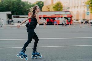 una foto al aire libre de una joven activa y delgada disfruta del patinaje sobre ruedas durante el tiempo libre vestida con poses de ropa negra activa en un lugar urbano en la carretera contra un fondo borroso con transporte. concepto de afición