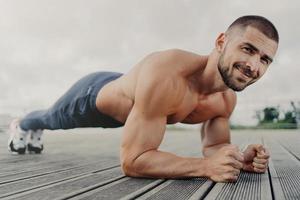 Workout and healthy lifestyle concept. Pleased muscular bearded young man stands in plank pose, makes sport training outdoors, looks with determined expression, concentrated on getting better photo