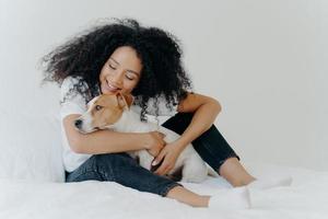 una mujer bonita con peinado rizado juega y disfruta con un adorable perro pequeño en la cama, tiene buen humor, posan juntos en un dormitorio acogedor. la mujer se despierta con su mejor amiga. adorable mascota con dueño en el interior foto