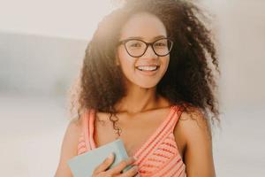 Photo of carefree African American student in summer dress, holds book, prepares for exam, strolls across city streets, enjoys warm sunny day, has charming smile on face. People, ethnicity concept