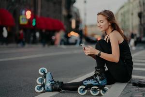 una foto al aire libre de una joven deportiva activa vestida con ropa activa usa patines revisa el contenido del mensaje en poses de teléfonos inteligentes contra el fondo borroso de la ciudad en el asfalto lleva un estilo de vida saludable