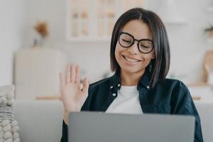 la chica emprendedora con gafas tiene una videollamada en la computadora y saluda con la mano. trabajo a distancia en casa. foto