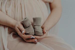 Maternity and pregnancy concept. Expectant mother prepares for giving birth to child holds boots in hands poses indoor being on third trimester photo