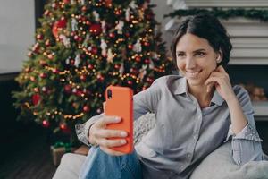 alegre joven española disfrutando de la Navidad en casa, haciendo selfie en smartphone foto