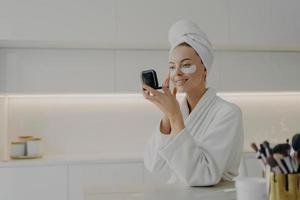 Happy woman in white bathrobe with towel on head taking care of her skin after morning shower photo