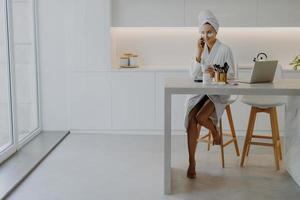 Pleased young woman dressed in bathrobe poses bare feet at chair in spacious kitchen sits at table with laptop and cosmetic products applies beauty patches under eyes talks via modern cellular photo