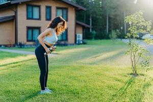 Full length shot of active sporty young woman raises dumbbells has workout outdoor near house stands on green lawn, dressed in sportsclothes, tries to be in good shape, stays fit and healthy photo