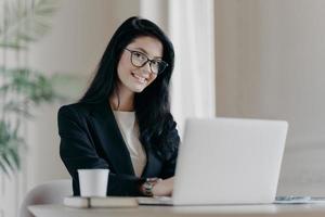 Positive dark haired woman makes research, browses information on laptop computer, connected to wireless internet, being happy wears formal clothes and spectacles drinks coffee. Economist works online photo