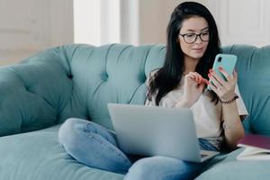 Serious brunette female freelancer works remotely on laptop computer, sends text messages via smartphone connected to wireless free internet sits crossed legs on comfortable sofa surfs social networks photo