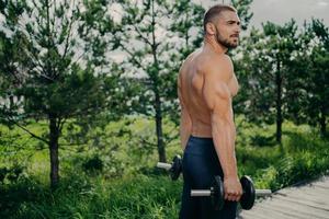 Outdoor shot of muscular strong man prepares for barbell workout, lifts weight and poses at street, gives motivation to everyone. Male weightlifter does biceps exercises, wears sportive clothes photo