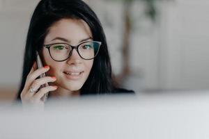 Horizontal shot of attractive brunette female employee with concentrated face expression, talks on mobile phone, has cellular conversation, wears spectacles for vision correction, busy working