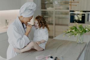 Happy European mother keeps cosmetic brush on daughters nose applies face powder dressed in white soft bath robe take care about appearance and skin. Family beauty wellbeing childhood motherhood photo