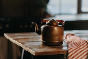Horizontal shot of aluminium teapot on wooden table against blurred background striped napkin near. Old kettle made of bronze