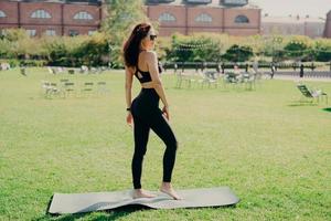 Fit brunette woman dressed in sportswear stands bare feet on karemat has fintess training on green lawn outdoor during summer time wears sunglasses looks somewhere being in good physical shape photo