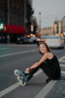 Outdoor shot of active slim woman poses on asphalt puts on rollerskates being in good mood spends free time riding rollerblades in urban place. Blurred city background. Hobby and recreation. photo