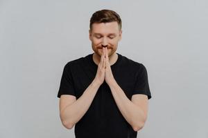 Young happy man in black t shirt with closed eyes keeping palms pressed together and dreaming about something photo