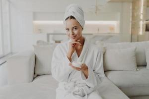 Young happy woman holding cosmetic face cream and smiling at camera while relaxing on sofa at home photo