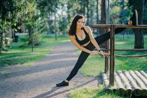 una joven flexible y rizada usa polainas y zapatillas negras, estira las piernas, hace deporte durante el verano al aire libre, posa contra árboles verdes. concepto de personas, flexibilidad y ejercicio. foto