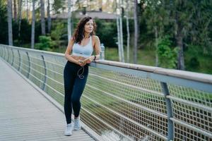 Serious thoughtful woman with spoty body, dressed in cropped top and leggings, poses at bridge looks pensively away, listens music in earphones. People, healthy lifestyle and fitness concept photo