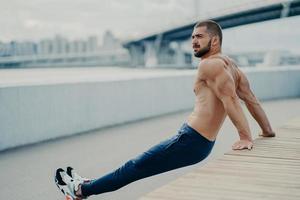 Purposeful bearded adult man trains biceps while doing reverse push up exercises poses with naked muscular torso concentrated away breathes fresh air, likes fitness and sport. Workout outside photo