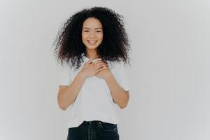 Photo of cheerful woman with Afro hair, keeps hands on chest, expresses gratitude, smiles gently, wears white t shirt and jeans, impressed to get pleasant compliment, isolated over white background