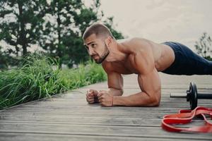 Muscular bodybuilder stands in plank pose demonstrates endurance has strong biceps poses outdoor has athletic workout, poses outdoor, keeps good physical shape. Man doing push ups, concentared forward photo