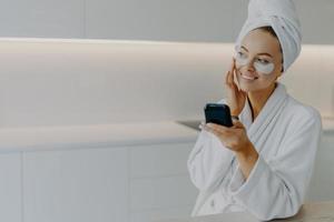 Happy smiling European woman applies moisturising patches under eyes touches face gently enjoys skin care routine dressed in white bathrobe looks somewhere aside poses at kitchen. Beauty concept photo