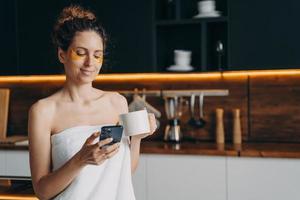 Happy girl applies collagen eye patches after shower and reading messages on mobile phone. photo