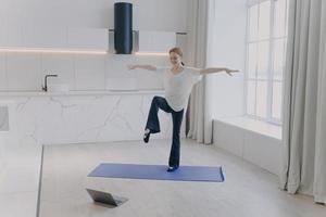 joven mujer feliz está haciendo ejercicio de equilibrio de pie sobre una pierna y estirando las manos en casa. foto