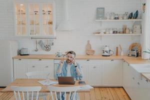 joven oficinista que trabaja de forma remota desde casa con una computadora mientras está sentado junto a la mesa de la cocina foto