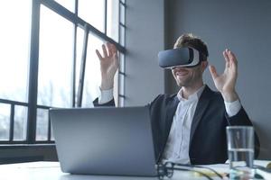 Young businessman in suit sitting at office table and using vr glasses goggles at work photo