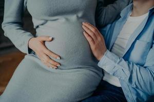 Pregnancy and family concept. Cropped shot of pregnant woman touches belly awaits for baby poses on husbands hands. Man touches stomach of wife anticipate child photo