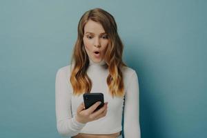 una chica milenaria sorprendida mira el teléfono móvil con la cara sorprendida, los ojos abiertos y la boca abierta foto