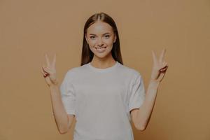 Una foto horizontal de una joven caucásica feliz y optimista levanta los brazos y muestra un gesto de paz con los dedos. El signo de la victoria dice que el número dos lleva una camiseta blanca informal aislada sobre un fondo marrón.