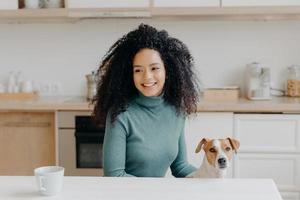 Happy dark skinned girl looks thoughtfully away, has pleasant smile, dressed in casual wear, drinks hot beverage, poses with domestic animal against kitchen interior. Female dog owner at home photo