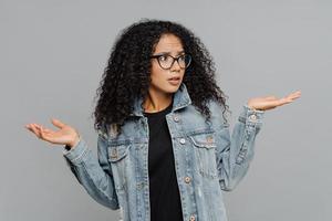 Confused unaware Afro woman with crisp hair, raises hands in bewilderment, looks aside, cannot make decision, wears spectacles and jean jacket, isolated over grey background. What should I do now photo