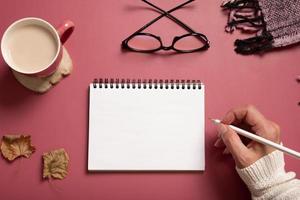 Table desk with notepad and female hand wit pencil and fall leaves on burgundy colored background. Autumn flat lay composition with copy space photo
