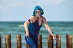 Artistic blue-haired woman performance artist smeared with blue gouache paints dancing on beach photo